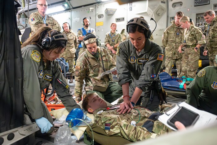 Tech. Sgt. Jenna Klingensmith performs CPR on a simulated patient aboard a C-17 Globemaster III while other medical personnel circle her to provide additional support with a group of observers in the background