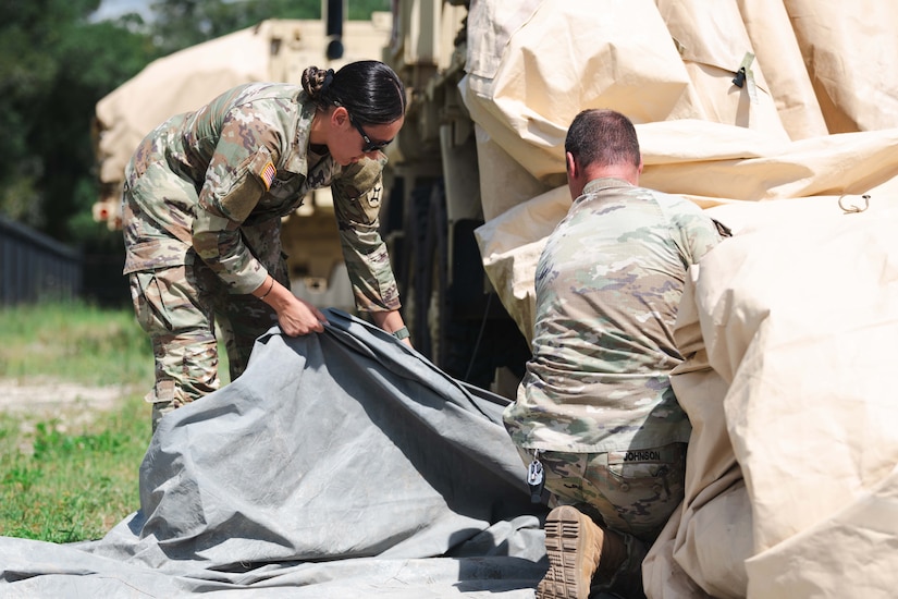 Two soldiers work to organize large canvas tarps near a military vehicle at daylight.