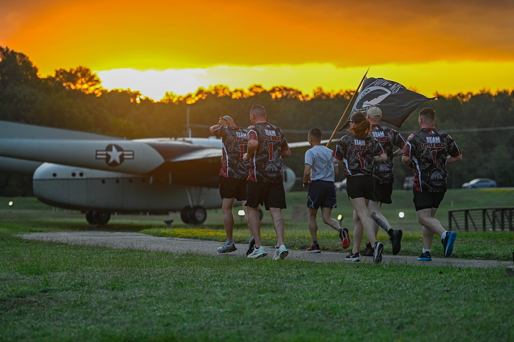 Men and women run in formation carrying a POW/MIA flag as the sun sets.