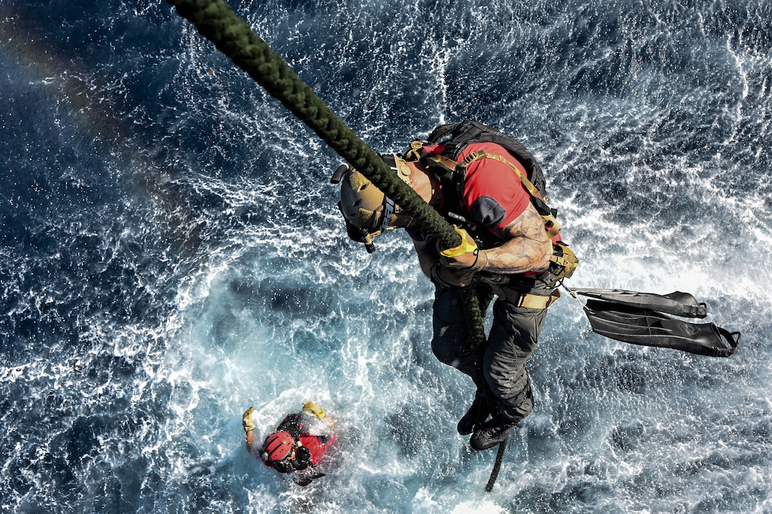 Overhead view of an airman fast-roping toward open water as another swims during daylight.