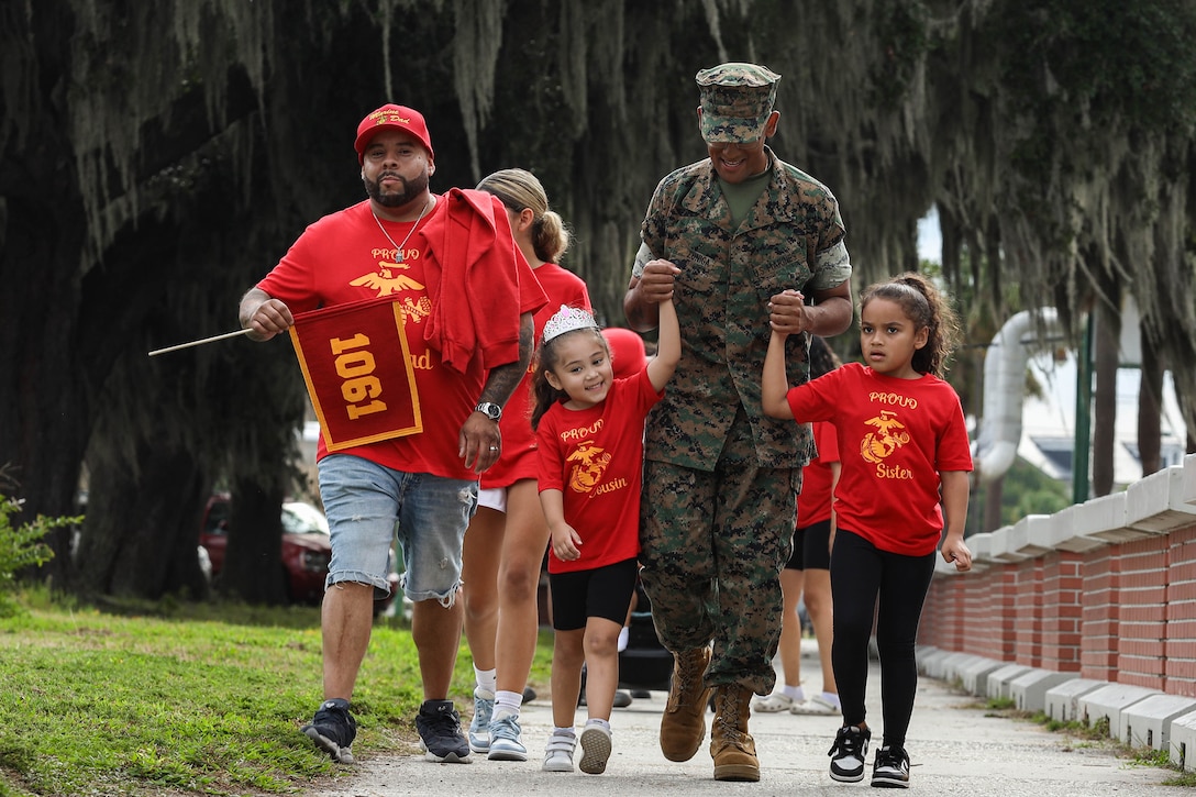 A Marine holds the hands of two children as other people walk in the background during daylight.