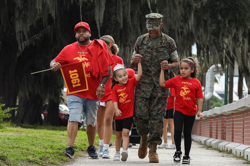 A Marine holds the hands of two children as other people walk in the background during daylight.