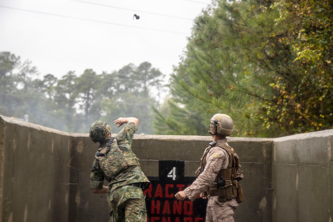 A Finnish service member throws a grenade toward power lines and trees while standing behind a concrete structure as another watches.