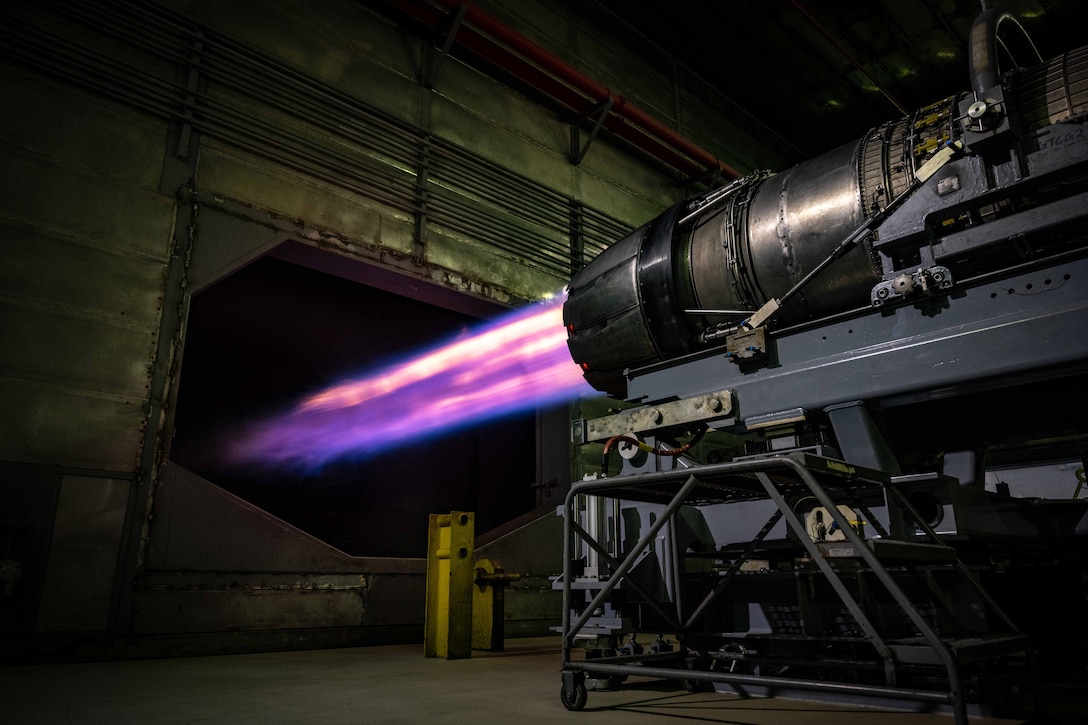 Purplish and orange flames come from a partially visible aircraft’s engine as it sits in a garage-like area.