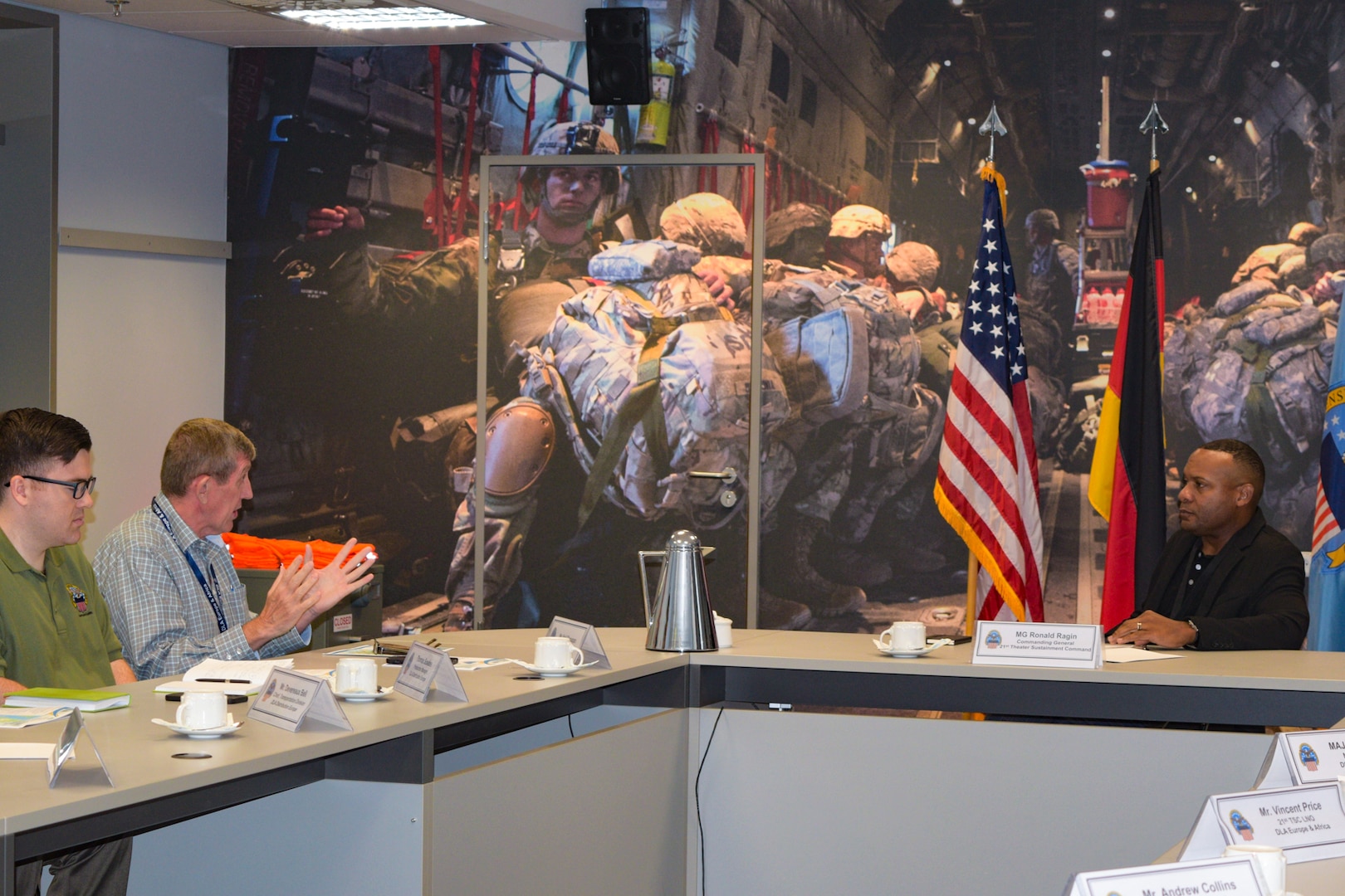 Three people sit at a long conference table talking. There are flags and military graphics on the wall behind them.