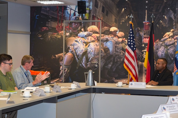 Three people sit at a long conference table talking. There are flags and military graphics on the wall behind them.