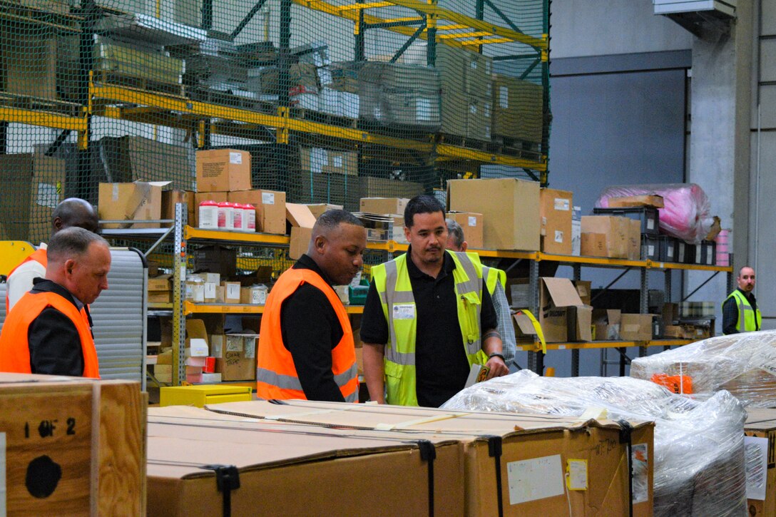 Four people in reflective vests look at a series of cardboard boxes in a distribution warehouse.
