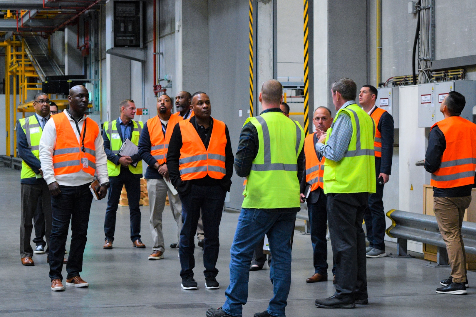 A large group of people wearing reflective vests stand talking in a distribution warehouse.