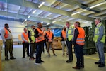 A group of people in reflective vests stand talking in a warehouse.