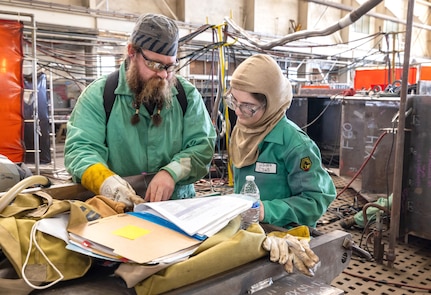 Welder Dan Carlson and welder apprentice Faith Chamness, Shop 26, Welders, look over a welding technique sheet before beginning work on a Reactor Compartment Disposal package support structure, Sept. 11, 2024, at Puget Sound Naval Shipyard & Intermediate Maintenance Facility in Bremerton, Washington. The cradle is being assembled inside Building 460 at PSNS & IMF. Shop 26's welder training career path can take someone with no welding experience and develop them into a highly skilled welder. (U.S. Navy photo by Wendy Hallmark)