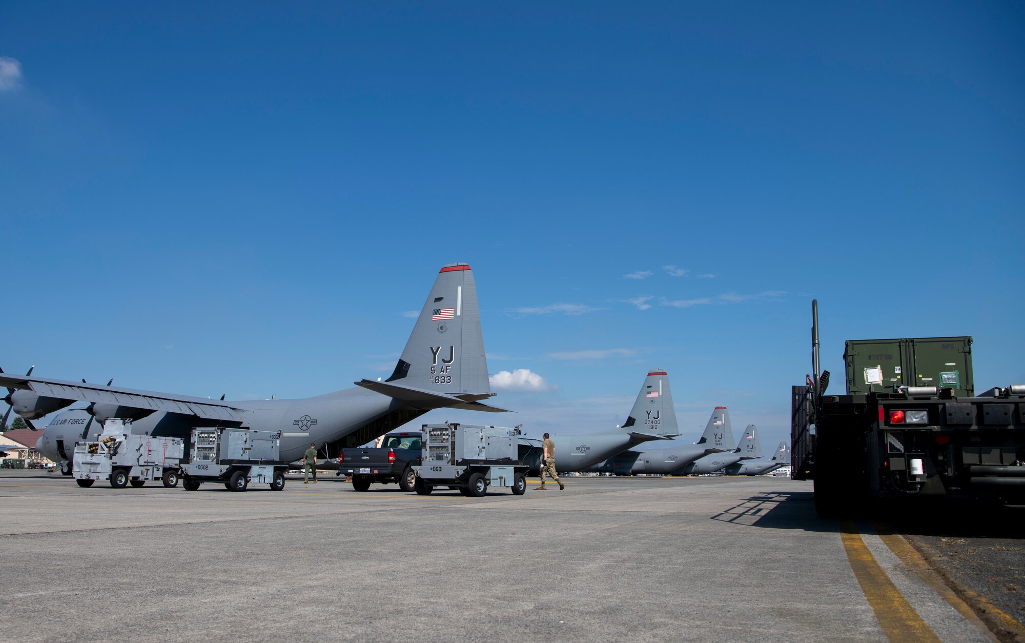 A row of C-130J Super Hercules are parked on the flight line, along with generators and a truck with a large green container on top.