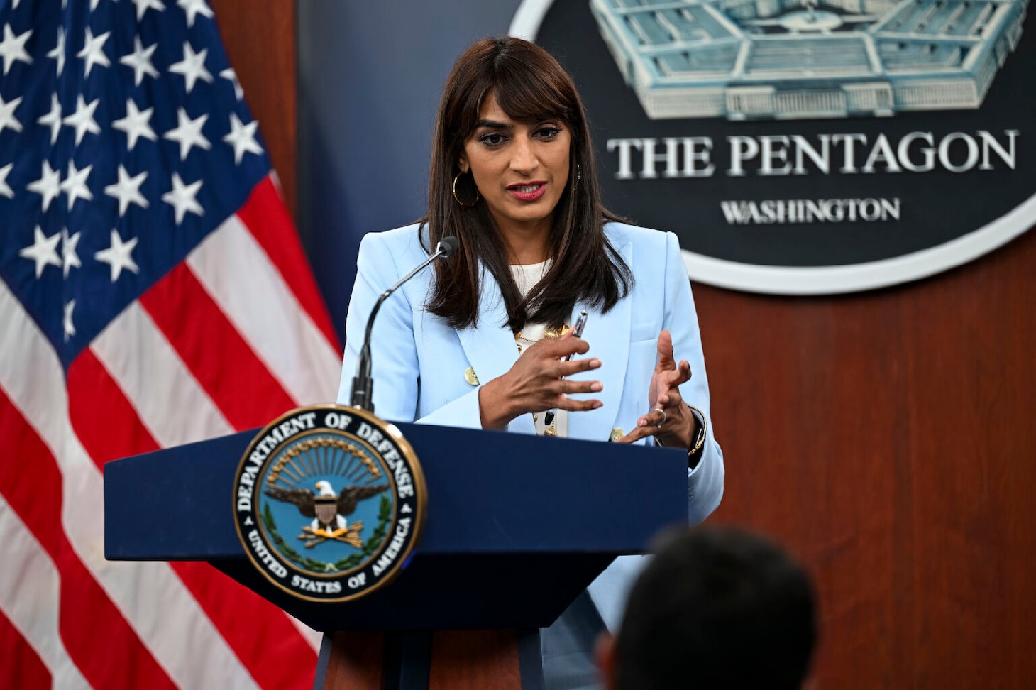 A person in business attire stands behind a lectern with an American flag in the background and a sign on the lectern that says, "The Pentagon - Washington."