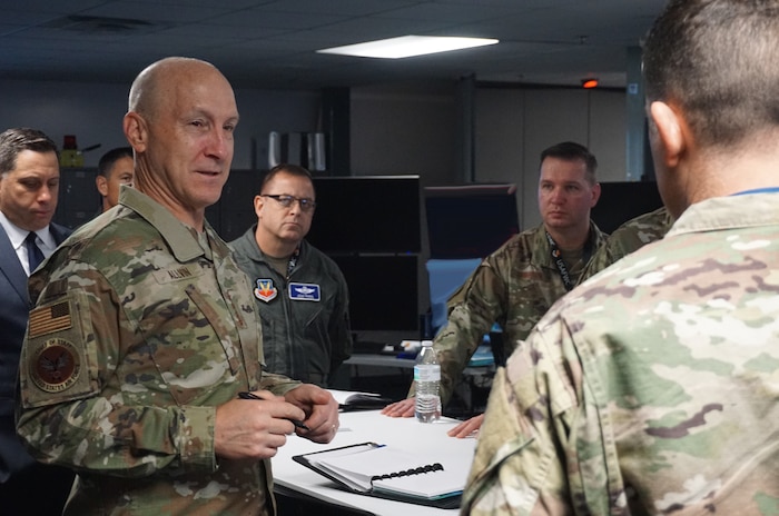 uniformed Chief of Staff of the Air Force Gen. David Allvin stands with uniformed U.S. military and civilians standing behind him and one in front of him briefing, with computer screens in the background