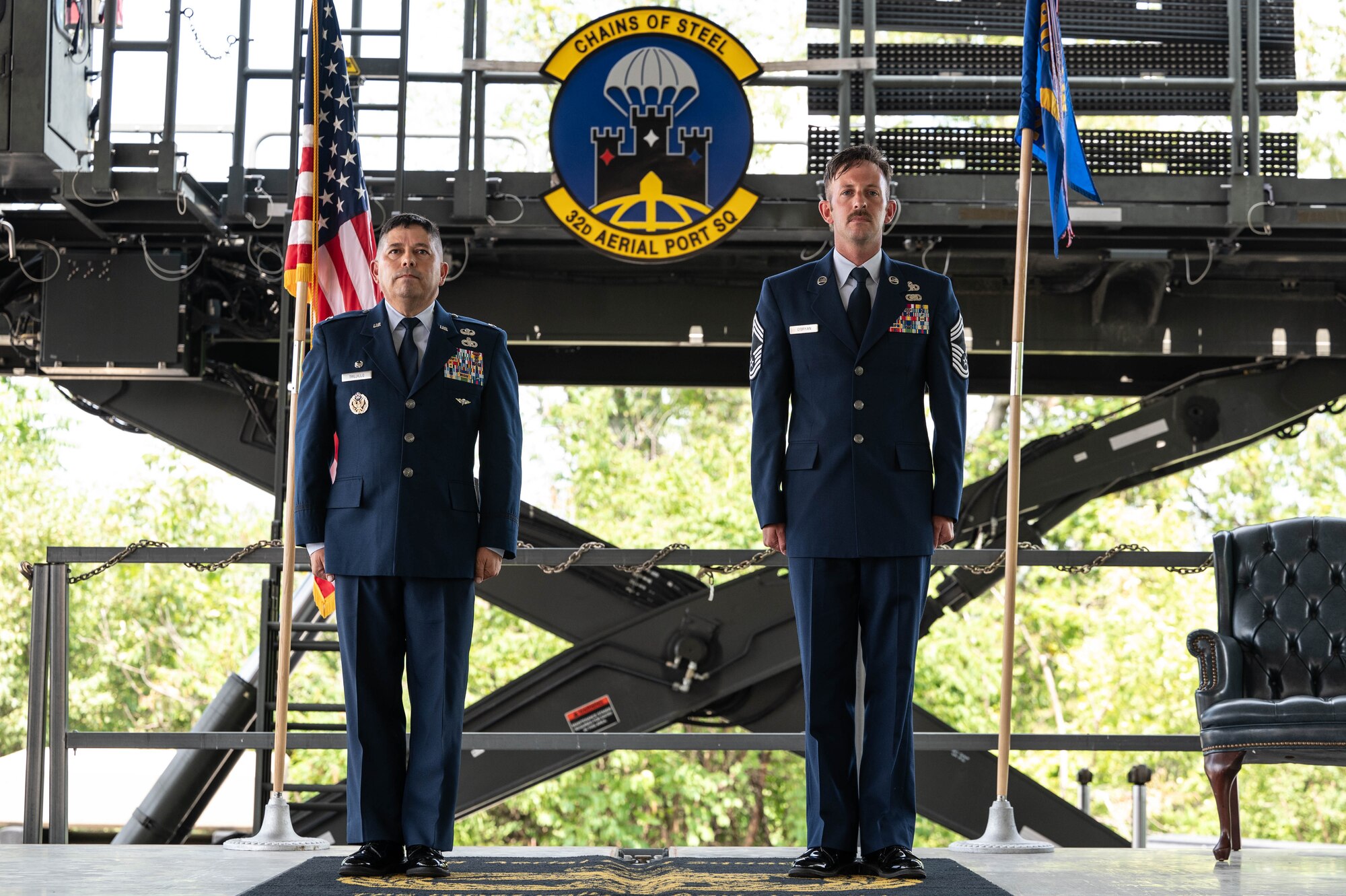 (From left) Col. Reginald Trujillo and Chief Master Sgt. Patrick O'Bryan stand at attention center of frame with the American flag and the 32nd Aerial Port Squadron flags on either side of them with a scissor lift bearing the 32nd APS insignia in the background