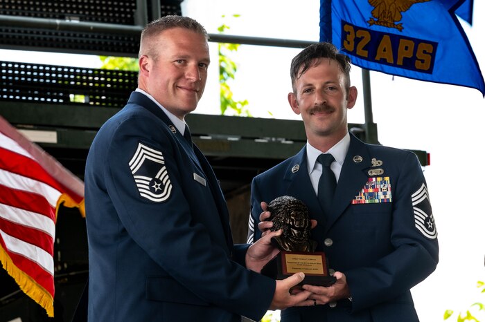 Chief Master Sgt. Chris Scott, left of screen, passes a chief's bust to Chief Master Sgt. Patrick O'Bryan as they both hold it and pose for the camera with the 32nd Aerial Port Squadron flag in the background