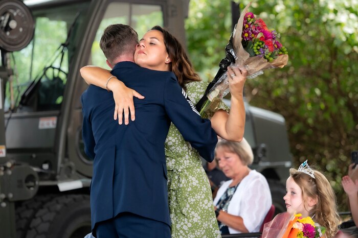 The back of CMSgt O'Bryan is to the camera as he hugs his wife with one arm wrapped around him and the other holding a bouquet of flowers with his family sitting behind them and a forklift in the background