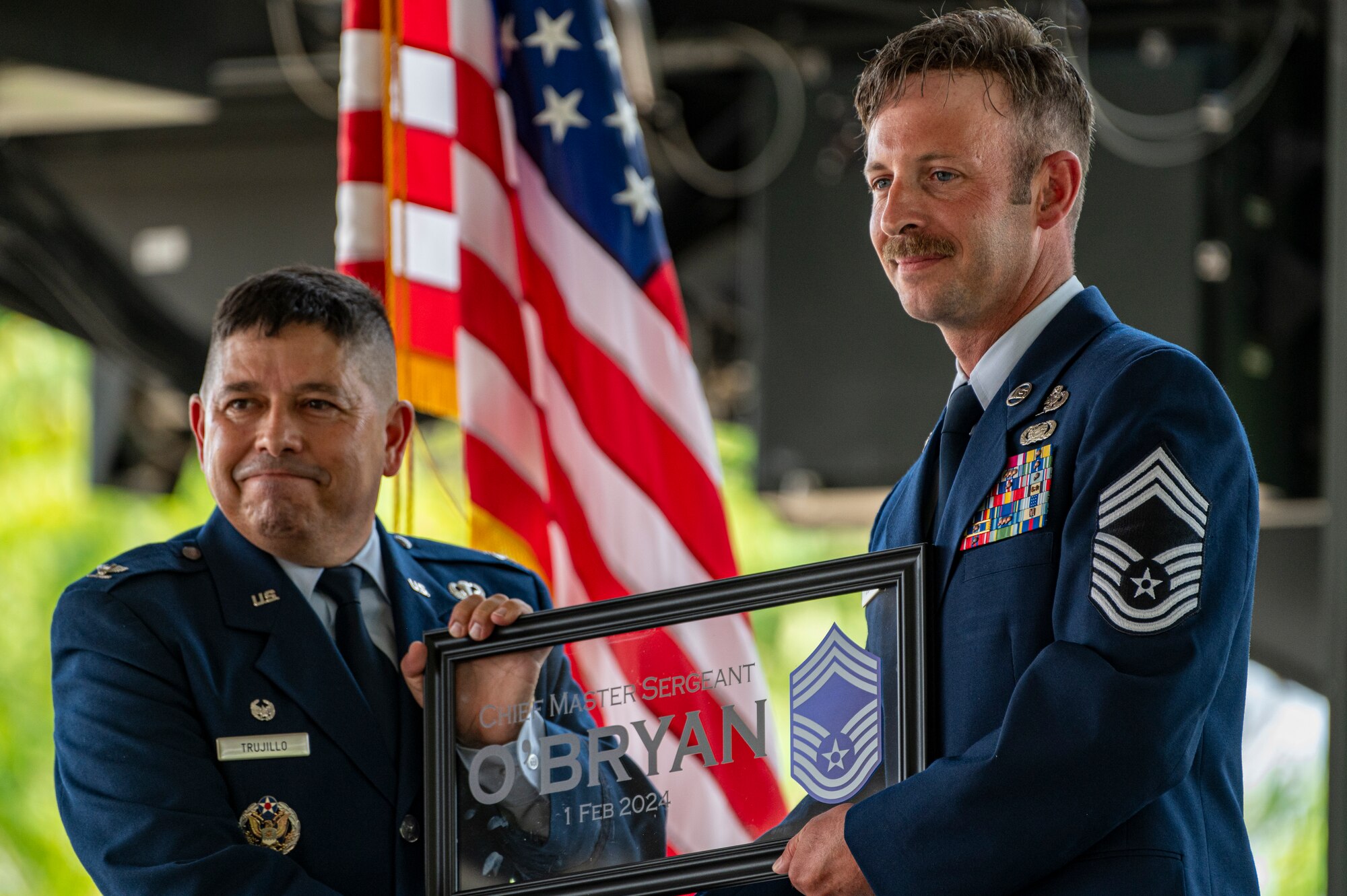 Col. Reginald Trujillo and CMSgt O'Bryan hold a gift presented to O'Bryan consisting of a transparent plaque etched and plastered with his name and rank as they pose for a camera out of frame and the American flag in the background