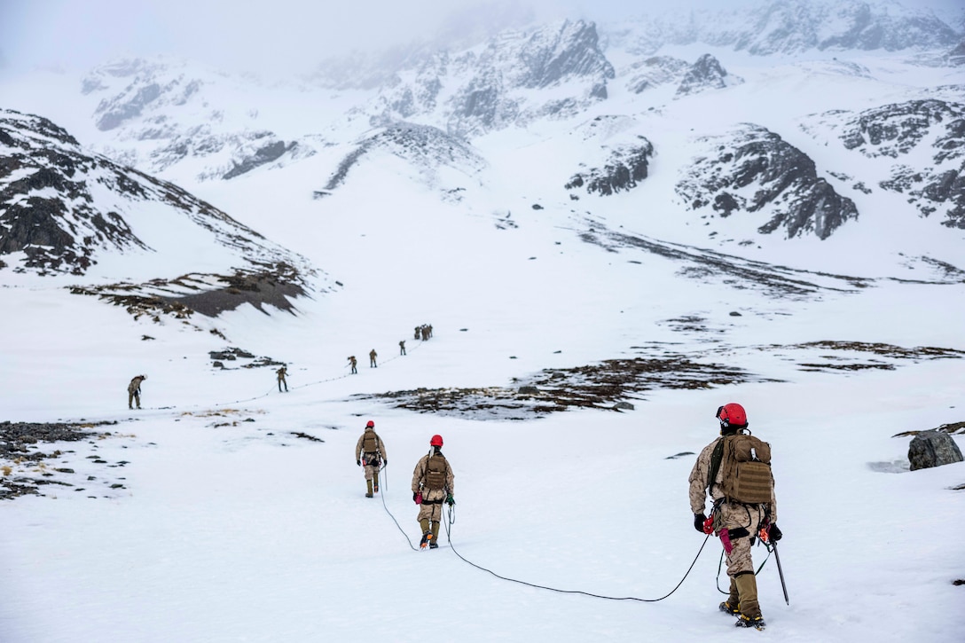 Marines using mountaineering gear hike up a snowy mountain pass with high peaks in the background.