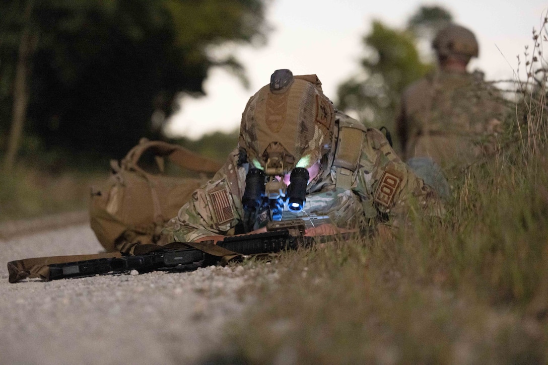 An airman wearing binoculars lies on the ground and looks closely at a device.