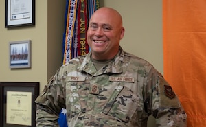 A bald man in a camouflaged uniform smiles for a photo. His name tapes read "Price" and "U.S. Air Force". In the background, an orange tapestry, a guidon with multiple streamers and various framed certificates and photos.