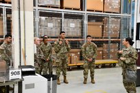 Several U.S. Army Soldiers stand in a warehouse, receiving a tour from another Soldier.