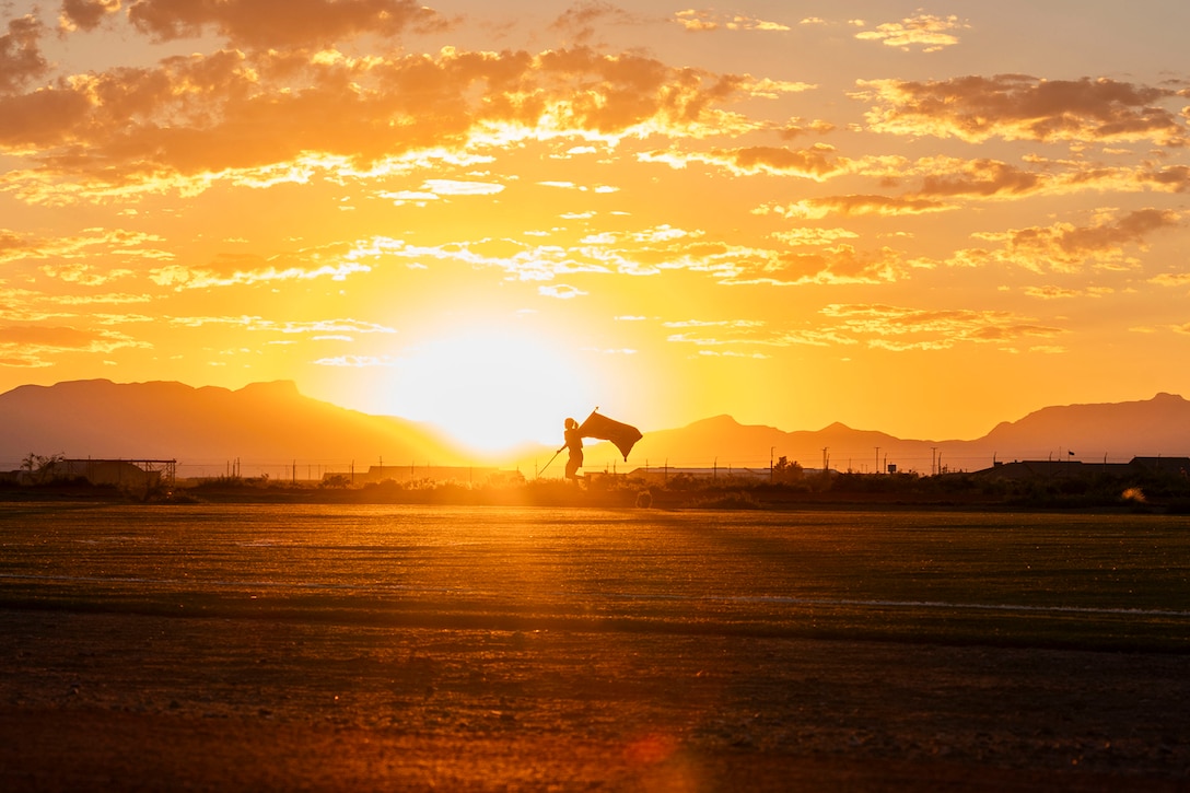 An airman, in silhouette, holds a flag while running with a low sun in the background.