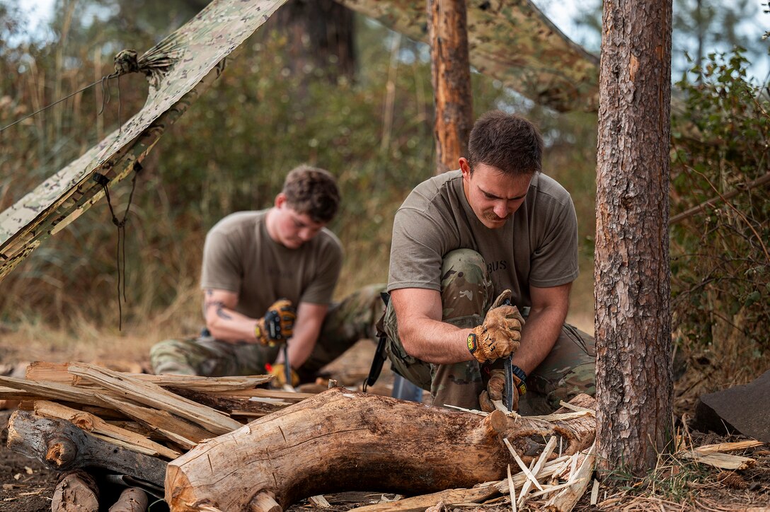 Two airmen kneel to split wood during training in a wooded area during daylight.