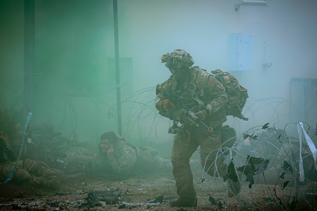 A soldier with a weapon runs near barbed wire through a bluish smoke as fellow soldiers take cover on the ground.