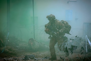 A soldier with a weapon runs near barbed wire through a bluish smoke as fellow soldiers take cover on the ground.