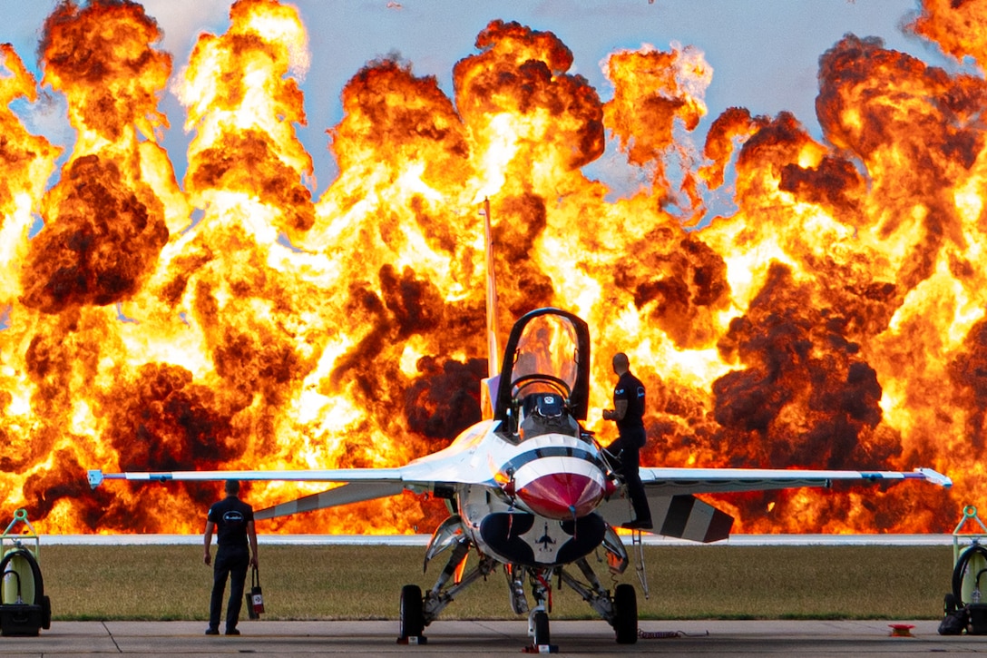 Two airmen standing on and near an aircraft look at a large explosion in the daylight.