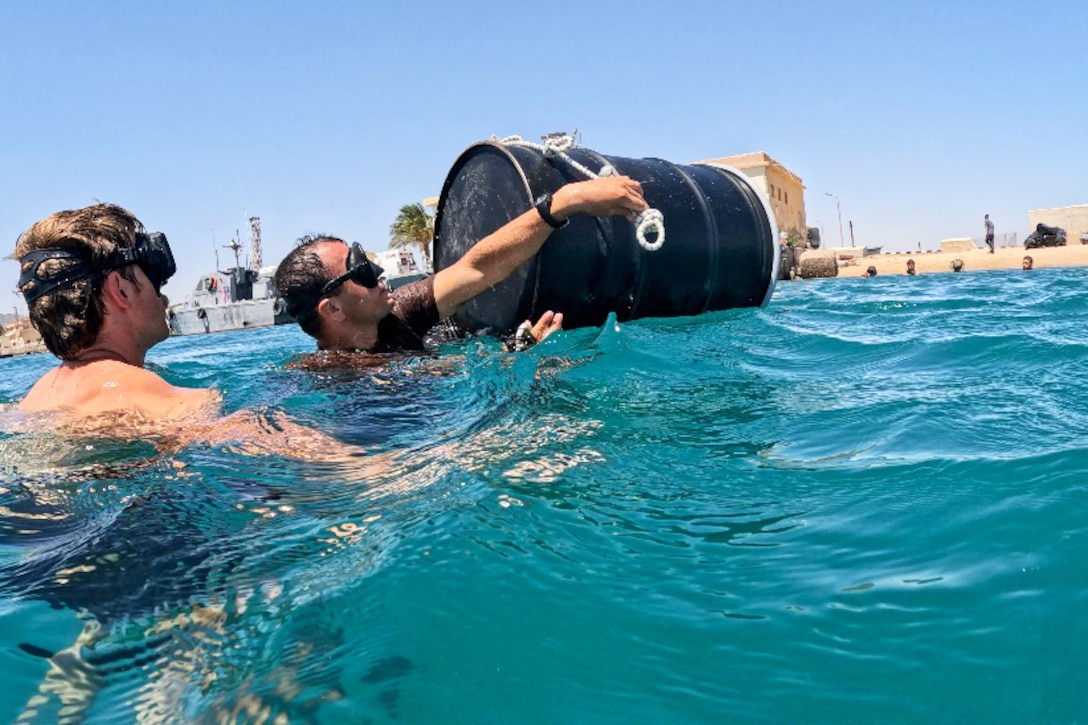 Two sailors wearing goggles swim near a barrel as fellow sailors swim near the shore in the background during daylight.