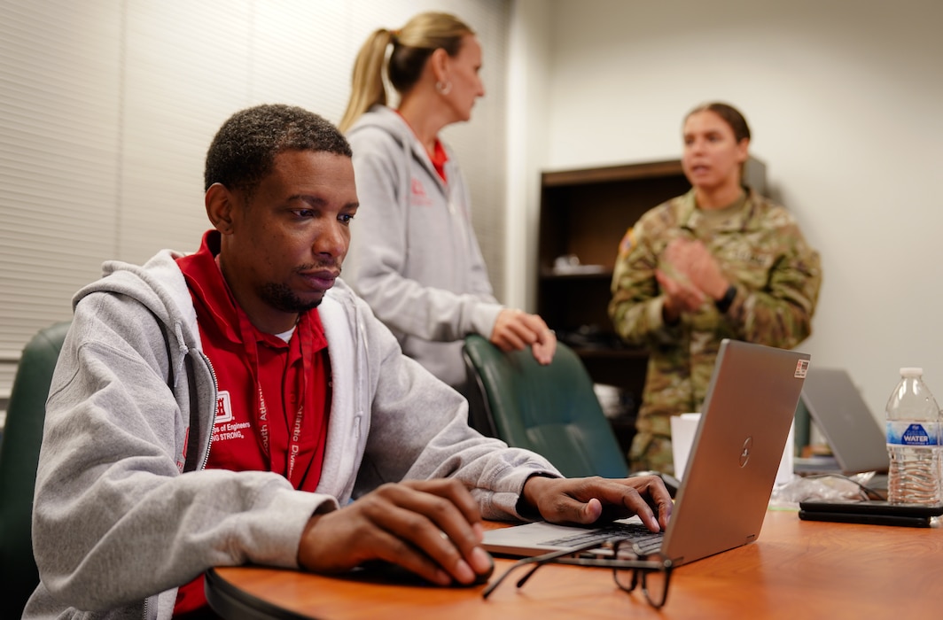 Emergency responder in a red U.S. Army Corps of Engineers shirt works at a computer with a civilian and uniformed Army member in the background.