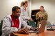 Emergency responder in a red U.S. Army Corps of Engineers shirt works at a computer with a civilian and uniformed Army member in the background.