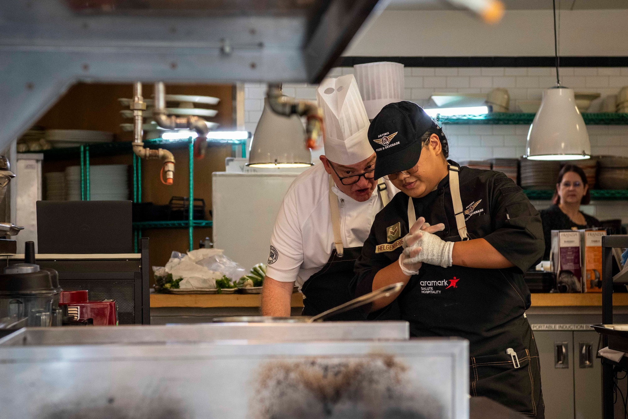 Chef Coach leans in to talk with Airman while she looks down at her dish in a pan on a stove