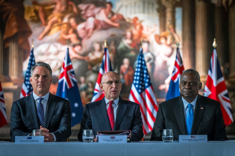 Three civilians sit next to each other at a long table.