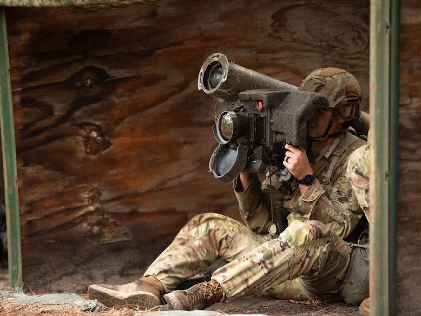 A soldier is seated in a wooden shelter and operates a shoulder-mounted weapons system.