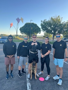 Four male Army Soldiers and one female Army Soldier dressed in Army PT uniform pose together with a German Shepherd