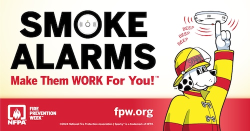 Image is of a dog dressed in a fire uniform pushing a smoke detector button.