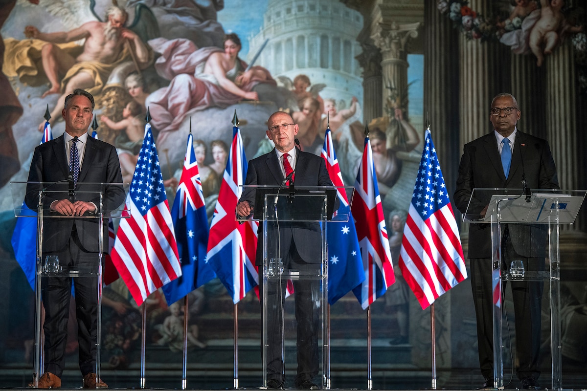 Three people in business attire stand behind lecterns and flags.