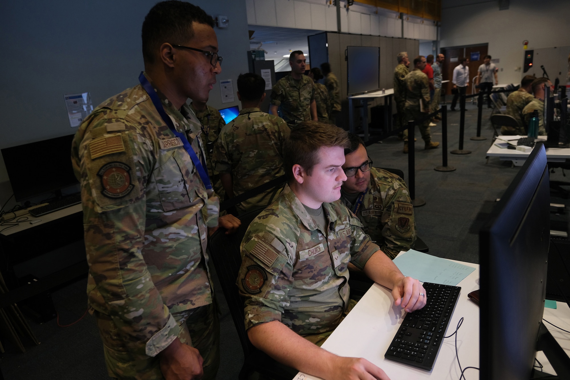 Uniformed US military members work on computers while other military and civilian members in the background are talking