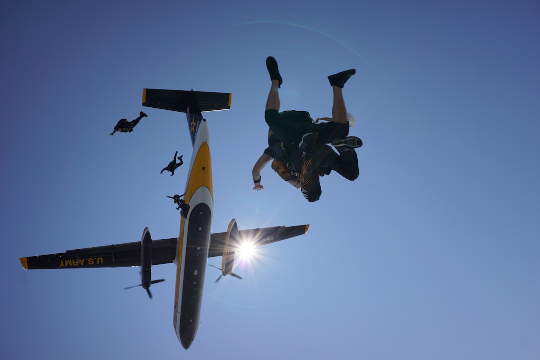 Soldiers freefall with parachutes under an aircraft as the sun beams nearby as seen from below.