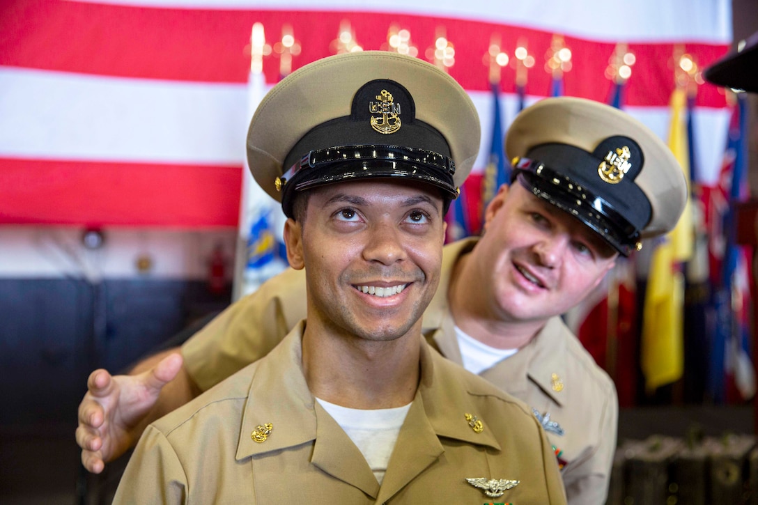 A seated sailor smiles as a fellow sailor watches from behind.