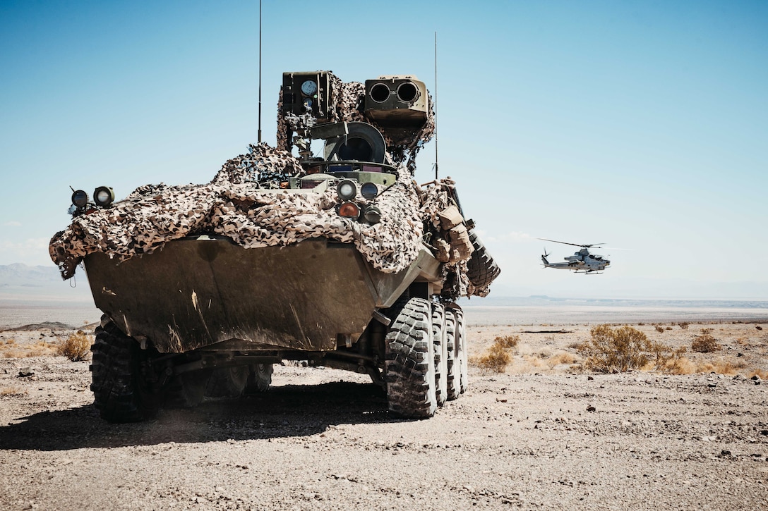 An armored vehicle sits in the desert as a helicopter hovers near the ground in the background.