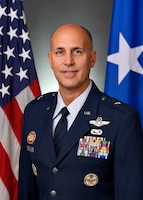 U.S Air Force general officer posing for an official portrait photo wearing Air Force service dress (blue suit and tie) in front of the U.S. flag and the blue one-star general officer flag.