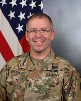 Army command sergeant major in combat uniform (light green camouflage) is posing for an official photo in front of a light grey background and the US flag.