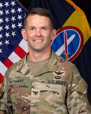 Army colonel in Army combat uniform posing for official photo in front of US flag and Military District of Washington command flag.