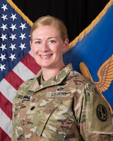 An Army lieutenant colonel in Army combat uniform (light green camouflage) is posing for an official photo in front of the U.S. flag and her command flag, which has a light blue background and a gold graphic image of wings over an airplane propeller.