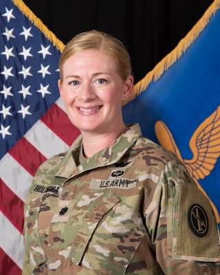 An Army lieutenant colonel in Army combat uniform (light green camouflage) is posing for an official photo in front of the U.S. flag and her command flag, which has a light blue background and a gold graphic image of wings over an airplane propeller.