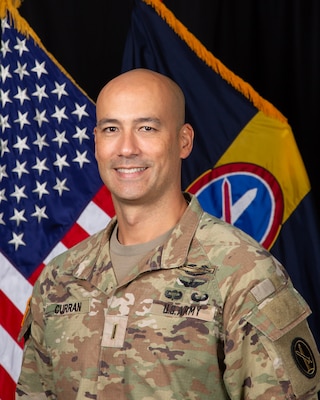 An Army chief warrant officer in Army combat uniform (light green camouflage) is posing for an official photo in front of the U.S. Flag and the command flag for the Military District of Washington.