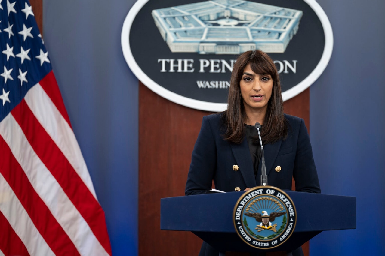 A woman in business attire stands at a lectern. The sign behind her indicates that she is at the Pentagon.